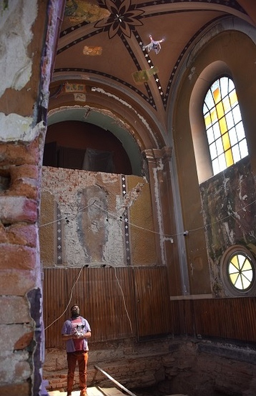 Fotografia wnętrza remontowanej kaplicy. W głębi widoczny jest operator sterujący dronem fotografującym z wysokości kilku metrów posadzkę oraz odsłonięte w trakcie badań archeologicznych relikty architektoniczne.